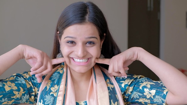 Closeup of a South Asian woman grinning at the camera while touching her cheeks with her fingers