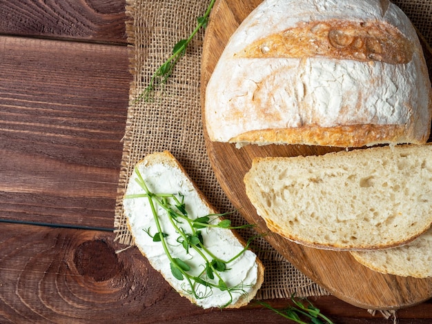 Closeup of a still life made of fresh crusty bread A slice of bread smeared with cottage cheese and decorated with micro greenery Linen napkin brown wooden background top view flat lay