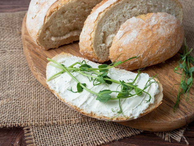 Closeup of a still life made of fresh crusty bread A slice of bread smeared with cottage cheese and decorated with micro greenery Linen napkin brown wooden background top view flat lay