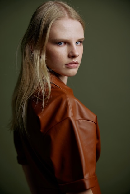 Photo closeup studio portrait of blonde with expressive glance blue eyes showy makeup looking over her shoulder at camera beautiful young woman in trendy leather brown dress posing isolated on green wall