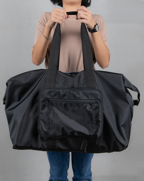 Photo closeup studio shot of female model in brown long sleeve shirt and jeans stand holding handle of fashionable trendy urban big gray unisex handbag with long shoulder strap in front of grey background.