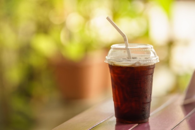 Closeup of takeaway plastic cup of iced black coffee Americano on wooden table with green nature background