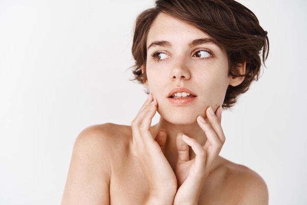 Closeup of tender young woman with naked shoulders gently toughing natural no makeup facial skin looking away pensive standing over white background