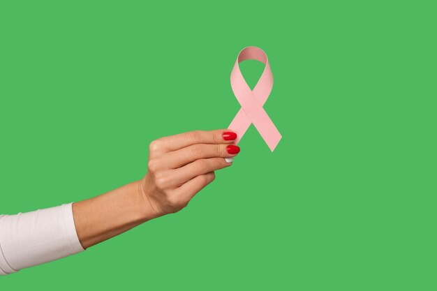 Closeup of tiny female hand with manicure holding pink ribbon, symbol of breast cancer awareness, oncological disease prevention and female healthcare. indoor studio shot isolated on green background