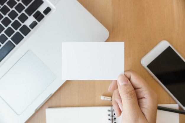 Closeup top view photo woman showing blank white business card and using modern laptop and mobile phone on wood table blurred background. Mockup ready for private information. Horizontal mock up.