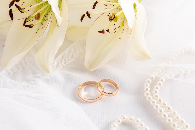 Closeup of two gold wedding rings and gorgeous white lily heads satin background with pearl beads design layout