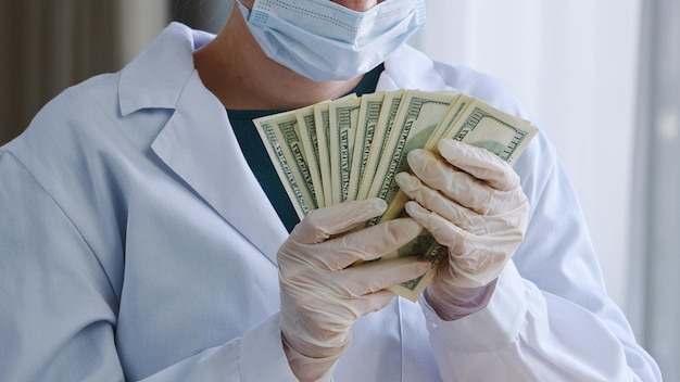 Closeup unrecognizable doctor nurse medical worker surgeon practitioner in white coat protective face mask and latex gloves holds stack of money counts bills banknotes receive bribe salary in clinic