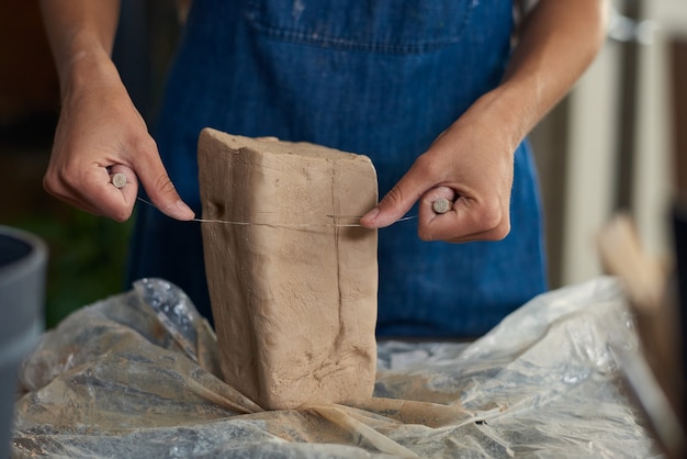 Closeup of unrecognizable potter in denim apron cutting clay with cutoff wire in workshop