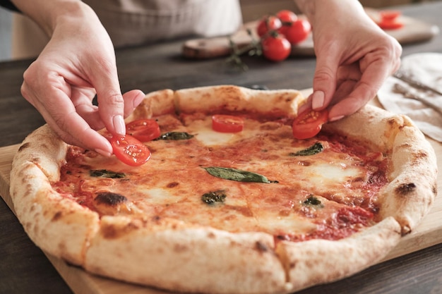 Closeup of unrecognizable woman decorating cheese pizza with tomato slices after baking