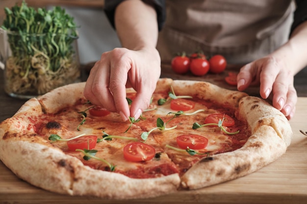 Closeup of unrecognizable woman putting green leaves on cheese pizza with cherry tomatoes while cooking tasty pizza