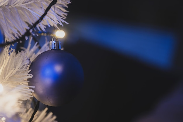 Closeup view of blue holiday bauble hanging on white christmas tree with shiny lights