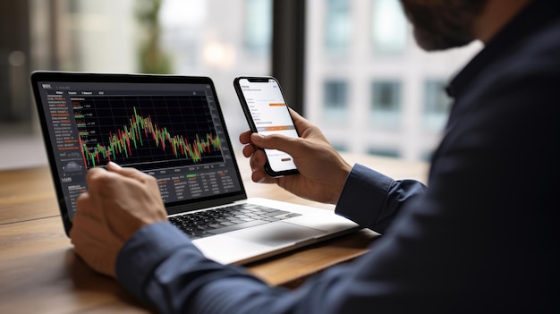 Photo closeup view of hands holding a smartphone with stock market data on the screen with a laptop in the background displaying financial charts