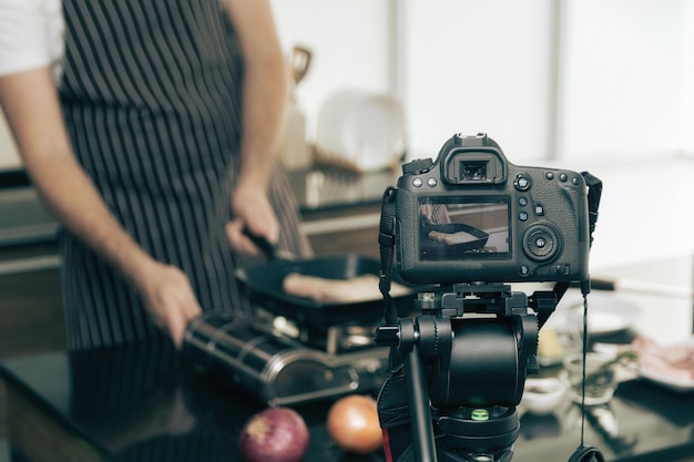 Closeup view of man in brown apron cooking at stove and shooting video of himself using DSLR camera on a tripod