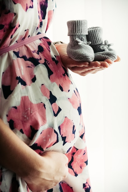 Closeup view of pregnant woman in floral printed dress holding small babys knitted shoes