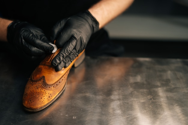 Photo closeup view of shoemaker wearing black latex gloves cleaning old light brown leather shoes with rag...