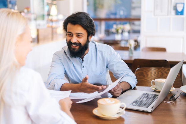 Closeup view of smiling Indian business man and Caucasian female business partners sitting together at table examining on documents