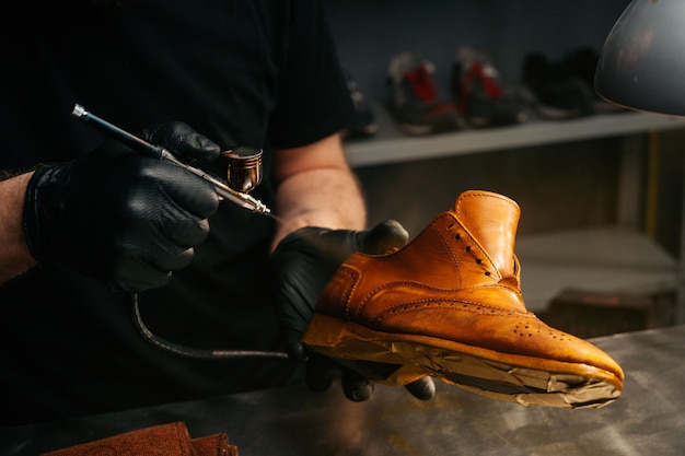 Closeup view of unrecognizable shoemaker wearing black gloves spraying paint of light brown leather shoes