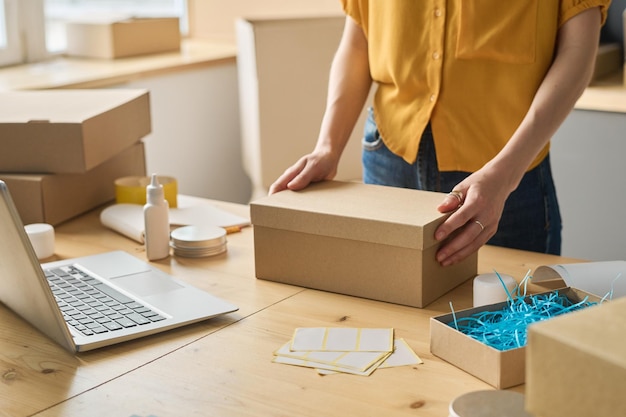 Closeup of warehouse worker packing online orders in cardboard boxes at table before shipping