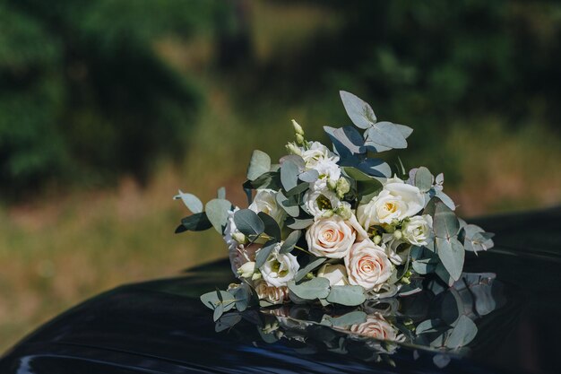 Closeup of a wedding bouquet of cream roses with green leaves lies on a black car