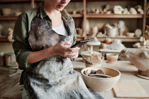 Closeup of woman in apron working online on her mobile phone while working with clay in the workshop