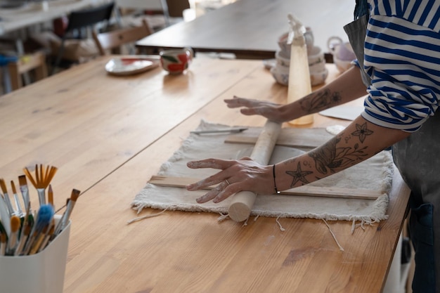 Closeup of woman ceramist in dirty apron rolling raw clay with the pin