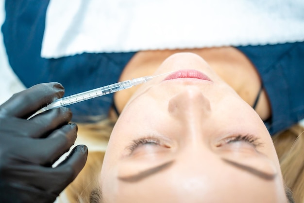 Closeup of woman face and hand in surgical glove holding syringe near her lips ready to receive beau