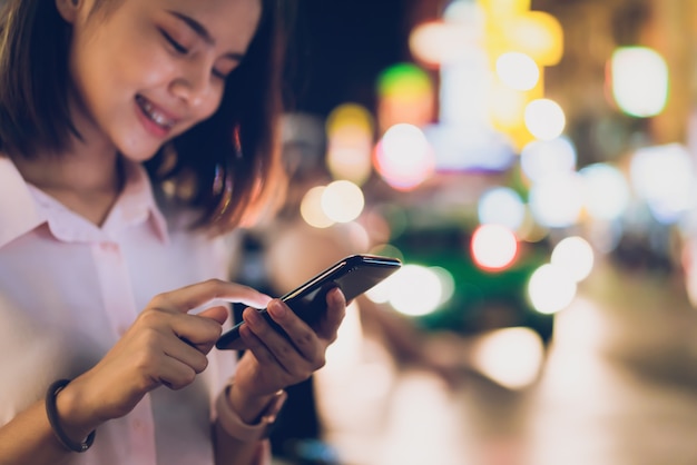 Closeup of woman holding a smartphone at night on city shopping street, and people walk. 