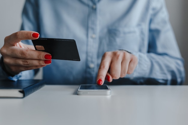 Closeup of woman's hands with red nails holding credit card and clicking mobile phone making payment online