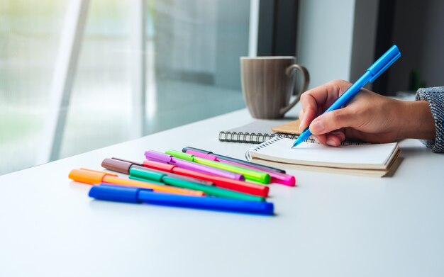 Closeup  of a woman writing on a blank notebook with colored pens and coffee cup on the table