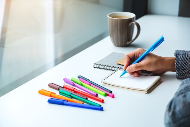 Closeup  of a woman writing on a blank notebook with colored pens and coffee cup on the table