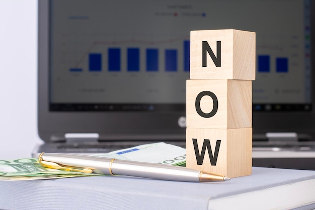 Closeup of wooden blocks with the word NOW and banknotes on the background of a laptop