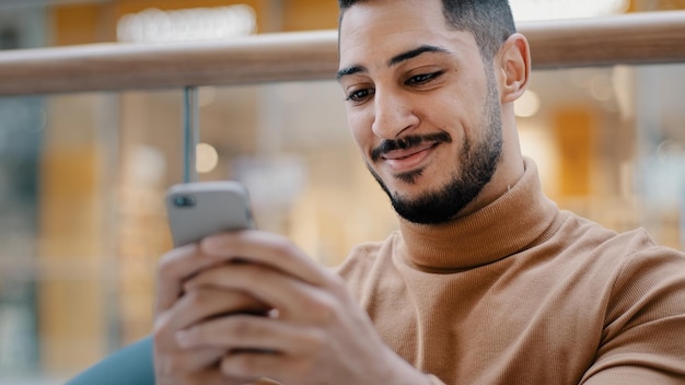 Closeup young arab guy holding phone looking at screen smartphone smiling receive nice email message