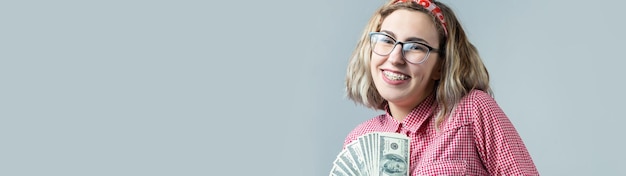 Closeup of young beautiful woman in a red plaid shirt in glasses with american dollars money in hand over gray background