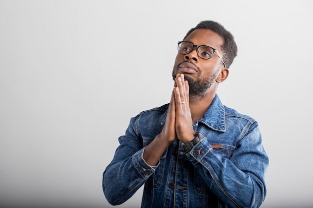 Closeup of young black man praying with hands clasped together, asking the God for good luck, success, health, looking up, isolated gray studio wall. Human emotion, facial expression, belief concept.