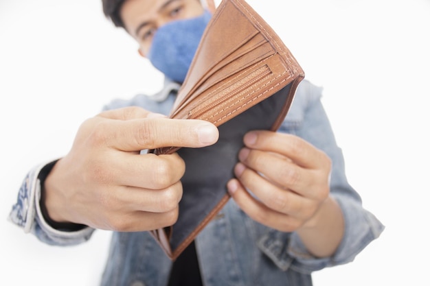 Closeup of a young man wearing a facemask, showing his empty wallet in front of a white background