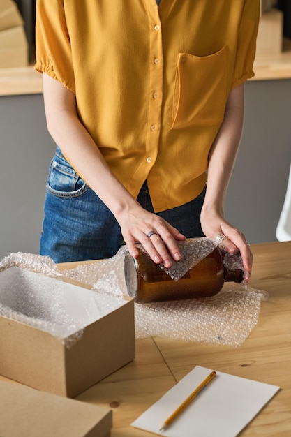 Closeup of young woman wrapping glass jar in packet she making parcel for shipping