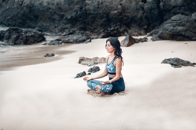 Closing her eyes, sporty lady meditates in lotus pose at the sandy beach by the rocks.