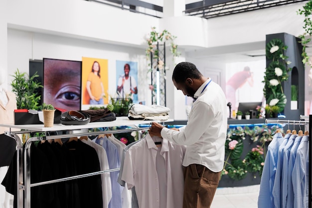 Clothing shopping mall man assistant hanging shirts on rack, displaying apparel for sale. Fashion boutique african american employee showcasing male garment for customers