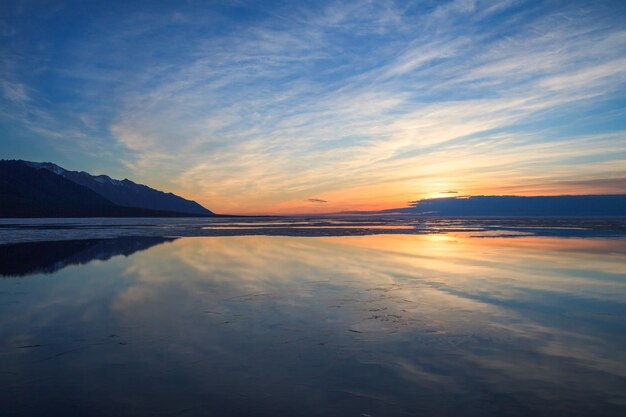 Cloudy sky and reflections in water surface with thin layer of ice on sunrise