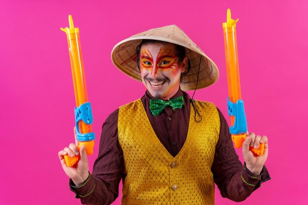 Clown with white facial makeup on a pink background smiling with toy guns