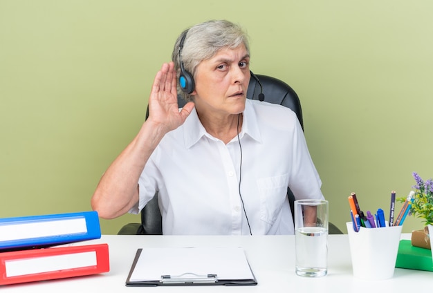 Clueless caucasian female call center operator on headphones sitting at desk with office tools keeping hand close to her ear trying to hear isolated on green wall