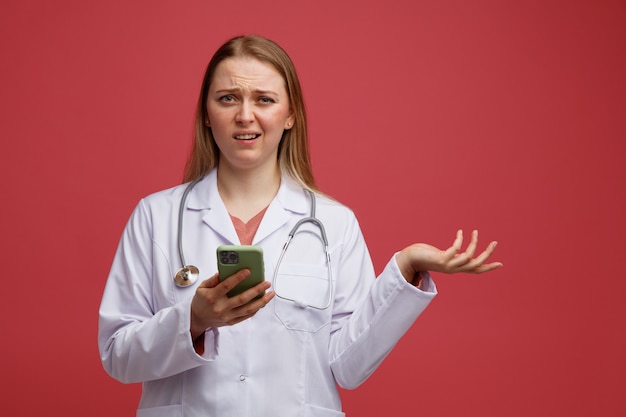 Clueless young blonde female doctor wearing medical robe and stethoscope around neck holding mobile phone showing empty hand 