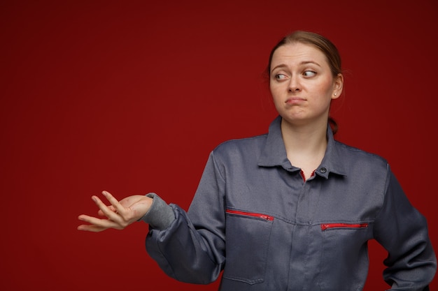 Clueless young blonde female engineer wearing uniform looking at side showing empty hand 