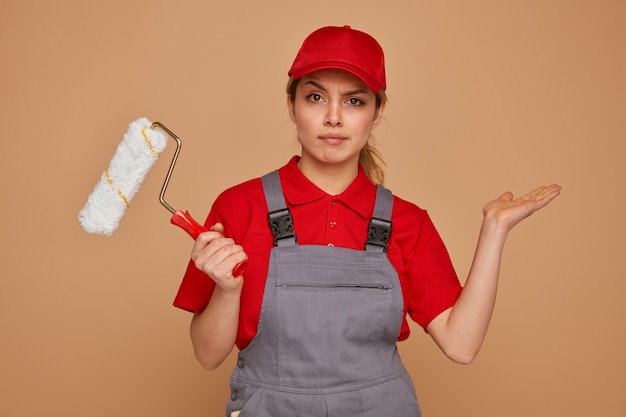 Clueless young female construction worker wearing cap and uniform holding paint roller showing empty hand 