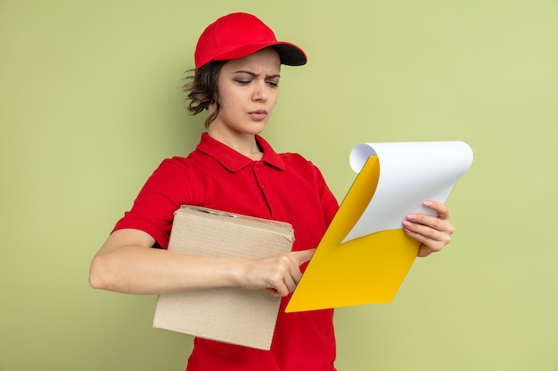 Clueless young pretty delivery woman holding cardboard box and looking at clipboard 