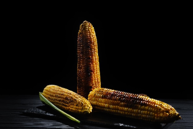 A cob of corn roasted over charcoal on a wooden board whole corn cobs dark background