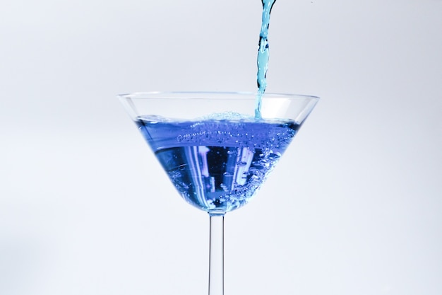 Cocktail with blue liquid in glass. Glass with blue water pouring with liquid with splashes and drops. Martini glass filling with alcohol with splashes on white background. Refreshing drink concept.