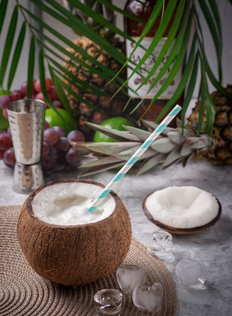 Coco loco cocktail on a bar table with ice cubes, coconut lid, some fruits in the background with palm leaves