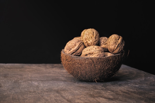 Coconut shell bowl full of walnuts in the shell on a wooden table on black wall