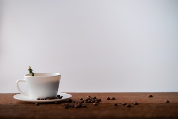 Coffee beans in white cup is on dark brown wooden table. A mini model of man is sitting on a cup.